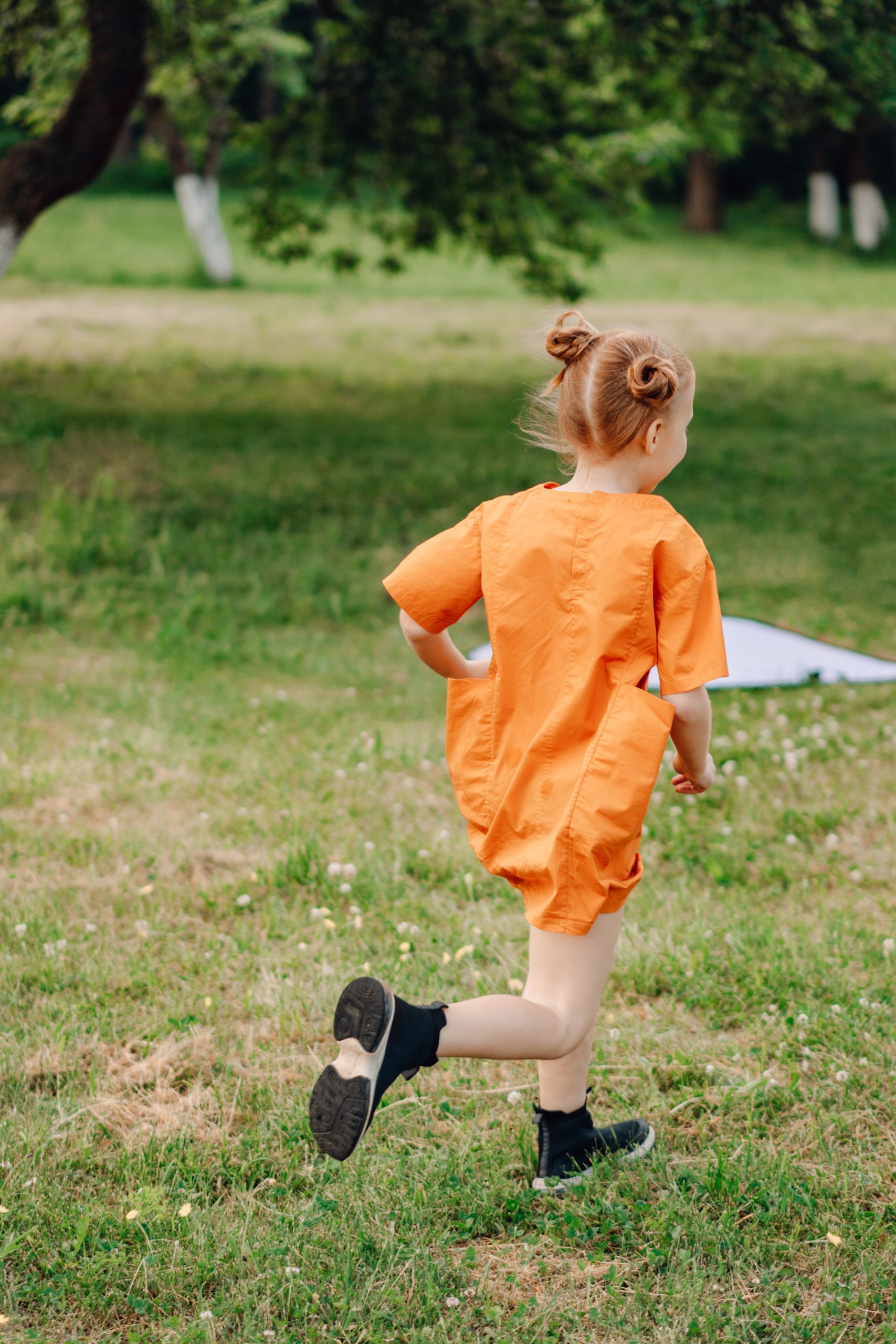 A joyful child with hair buns running through a grassy field in an orange dress, full of motion and energy.