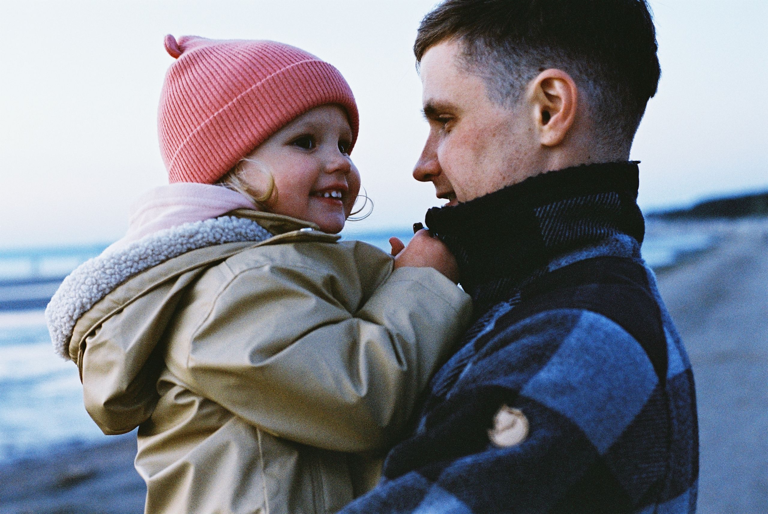 Father holds daughter, both smiling warmly by a serene St. Petersburg beach in winter.