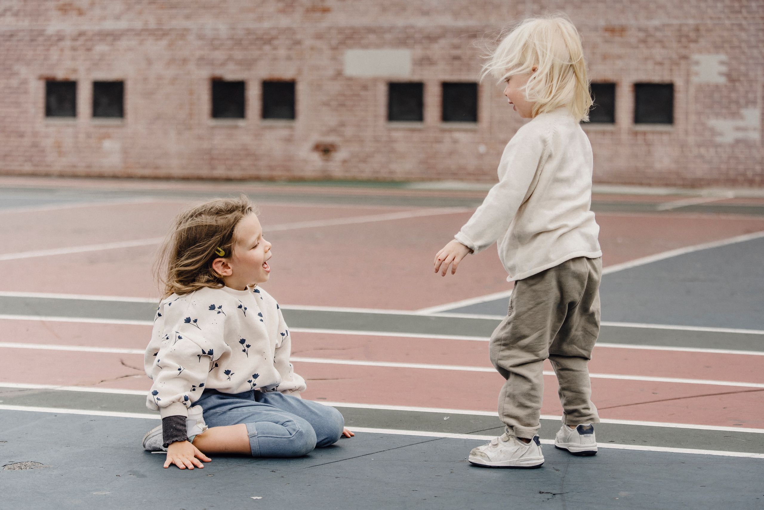 Side view full body of cheerful girl sitting near little boy standing on sports ground against brick wall on blurred background